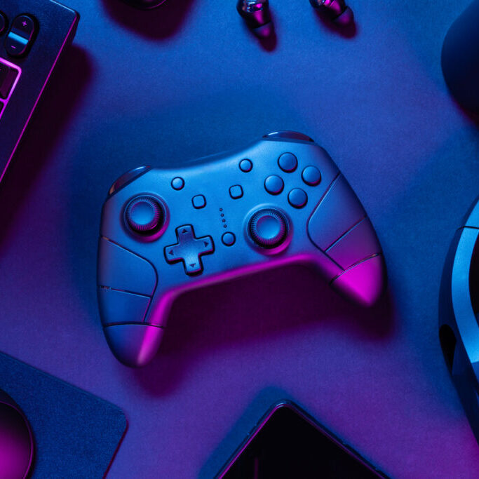 Overhead view of game controller with purple lit keyboard amidst various wireless devices and drink can on black desk